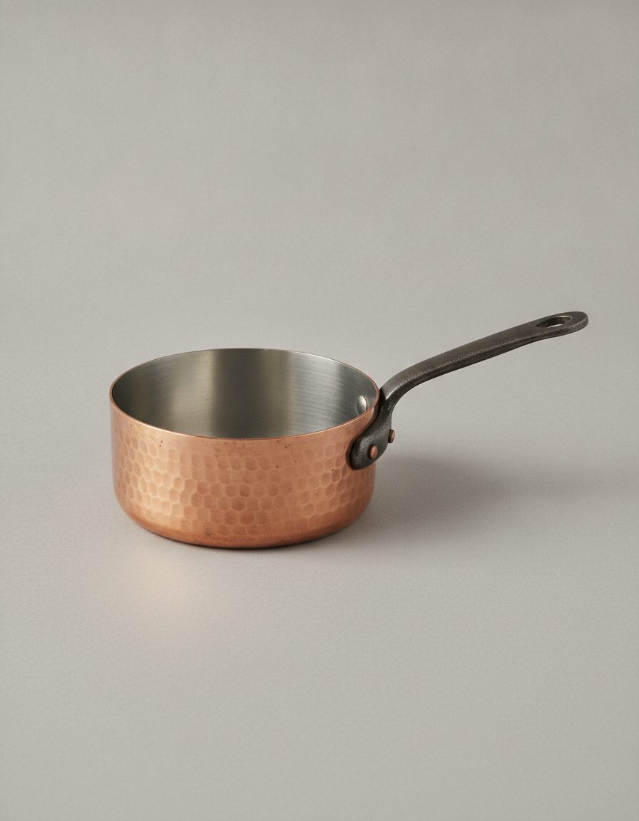A tin-lined copper saucepan with an iron handle photographed on a flat putty-grey background